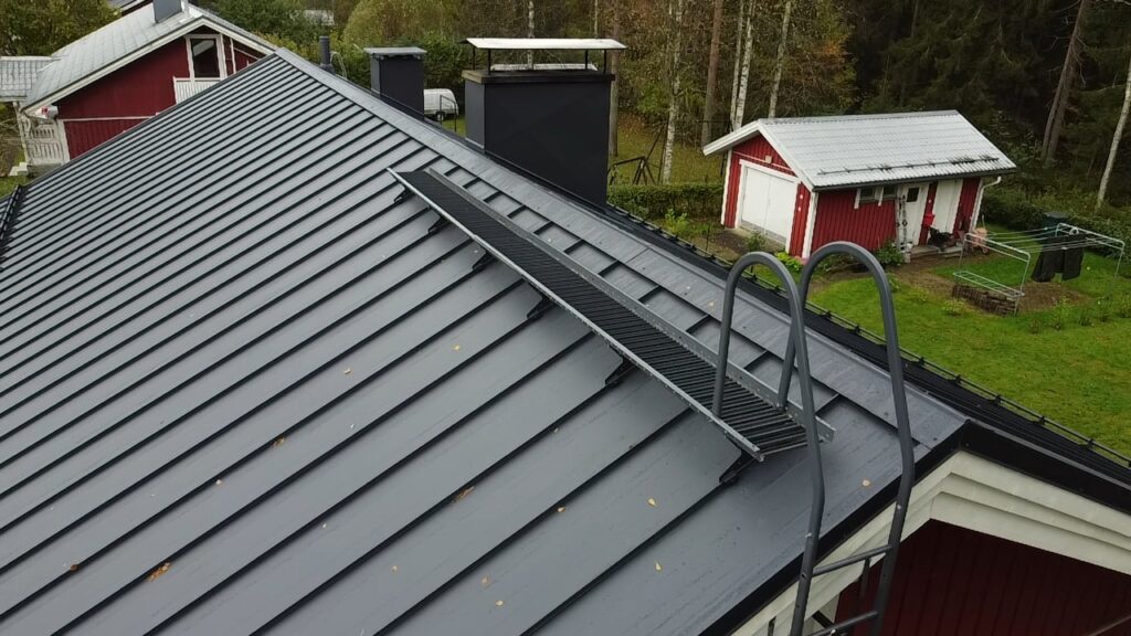 Ulkokuva omakotitalon harmaasta konesaumakatosta, jossa on mustat kattoturvatuotteet, kuten kattosilta, lapetikkaat ja lumiesteet.